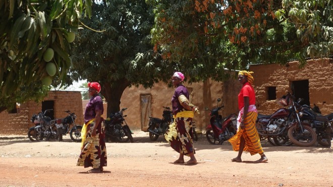 Drei Frauen in traditionellen Kleidern vor einem Mangobaum in einem Dorf in Burkina Faso. 